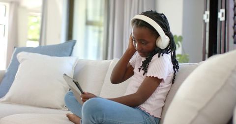 African american girl listening to music on headphones while using tablet on sunlit sofa