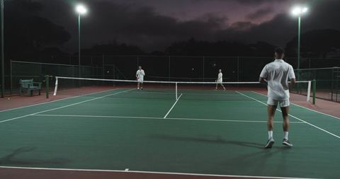 Night tennis match with diverse male players under floodlights on outdoor court at dusk