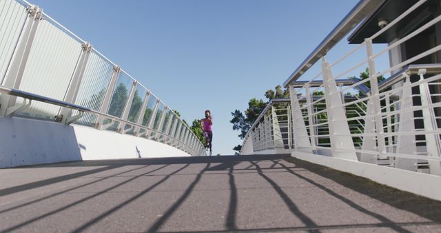 Woman running on a paved bridge in an urban area during a sunny day. Ideal for illustrating themes of health, fitness, and active lifestyle. Can be used in advertisements for sportswear, fitness apps, and urban development campaigns focusing on green cities and active living.