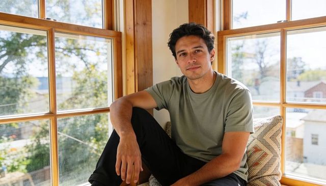 Young man relaxing on sunlit window seat in cozy home nook wearing olive t-shirt