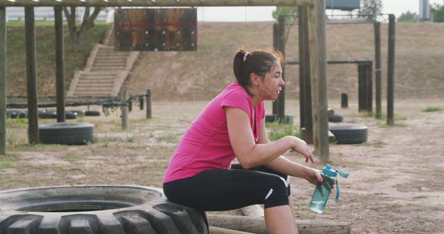 Woman in mid-workout attire sitting on a tire with a water bottle, taking a break during an intense outdoor obstacle course session. Suitable for themes around fitness, endurance training, outdoor exercise motivation, and hydration.