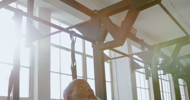 Senior man performing exercises with suspension trainer in sunlit gym setting. Ideal for content related to fitness, health, wellness, and active lifestyle for older adults. Useful for marketing fitness programs or products targeting senior populations.