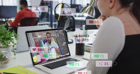 Woman watching live design tutorial on laptop with social media engagement overlays in open office