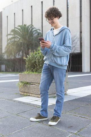 Teen smiling while checking smartphone in urban courtyard wearing hoodie and jeans