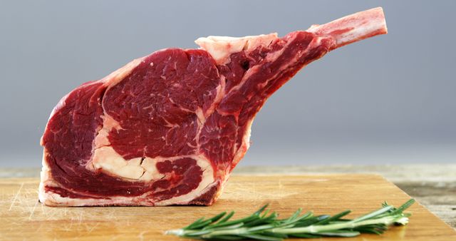 A raw, thick-cut ribeye steak with a prominent bone is displayed on a wooden cutting board, accompanied by fresh rosemary, with copy space. Its marbling suggests a rich flavor, ideal for grilling or roasting.