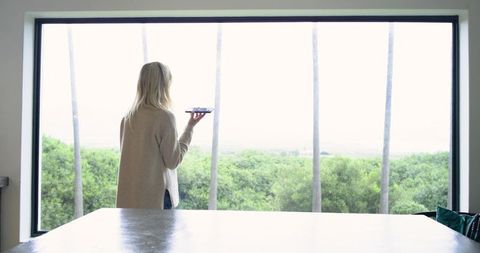 Woman holding smartphone at large window overlooking green palms and coastal panorama