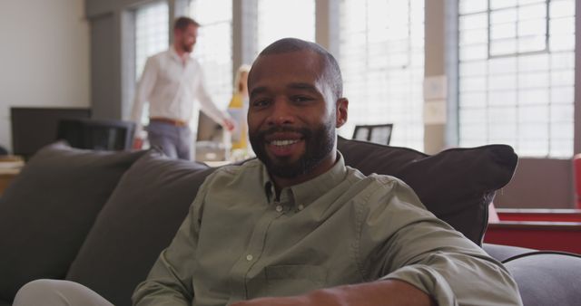 Man with beard smiling while sitting casually on a couch in a modern office. Ideal for illustrating workplace satisfaction, employee rest areas, modern office environments, and professional lifestyle. Can be used in business presentations, employee wellness programs, or workplace culture campaigns.