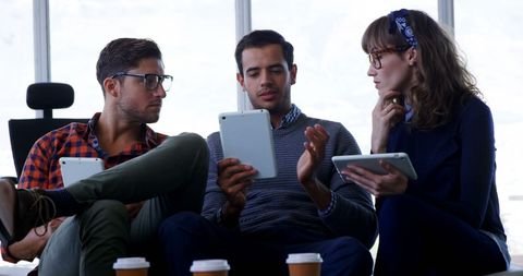 Group of young professionals engaged in conversation, exchanging ideas and utilizing tablets in a corporate setting. Highlights teamwork, modern technology, and effective communication. Useful for illustrating workplace collaboration, digital transformation in business, and productivity themes.