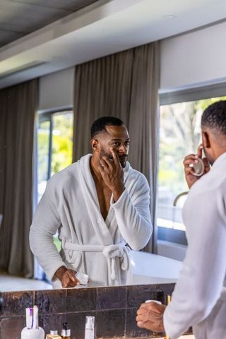 Mature african american man applying face cream at vanity mirror in white bathrobe