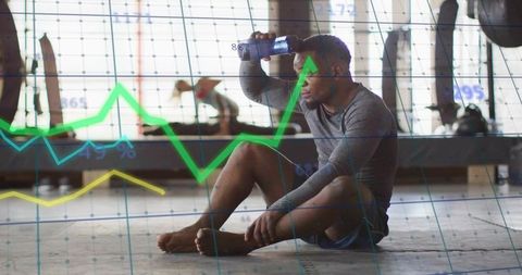 Male athlete resting on gym floor pressing water bottle to forehead with graph overlay