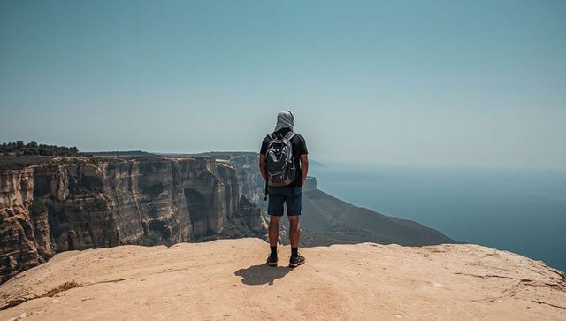 Solo hiker standing on coastal cliff overlooking vast ocean horizon, backpacking adventure