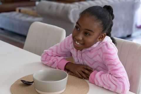 Smiling african american girl wearing pink fleece sitting at dining table with bowl