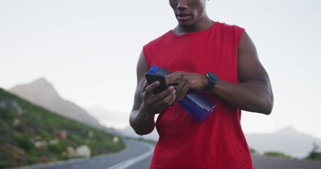 African american man holding water bottle using smartphone on the road. fitness sports and healthy lifestyle concept