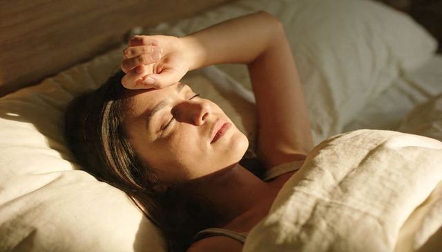 Sunlit bedroom portrait of relaxed woman resting under cream blanket in warm morning light