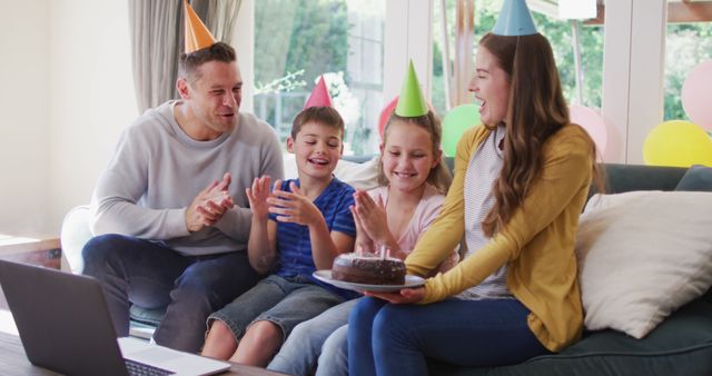 Family of four celebrating a birthday at home with a virtual party. Parents and children wear party hats and smile as they gather around a chocolate birthday cake. This image captures warmth, joy, and togetherness, perfect for themes related to family celebrations, virtual events, and happy moments. Ideal for use in blog posts, advertisements, or articles about family life, virtual birthdays, or special occasions.