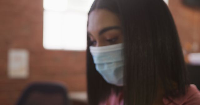 Young woman wears a protective mask indoors, highlighting pandemic safety measures. Blurred background suggests she could be in a variety of environments, such as a workplace, school, or home. Ideal for illustrating themes related to health, safety, pandemic, and responsible behavior during Covid-19.