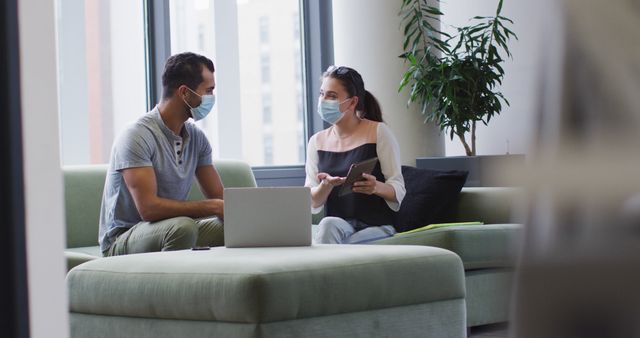 Diverse male and female business colleagues wearing masks sitting on sofa making elbow bump. working at the office of an independent creative business during covid 19 coronavirus pandemic.