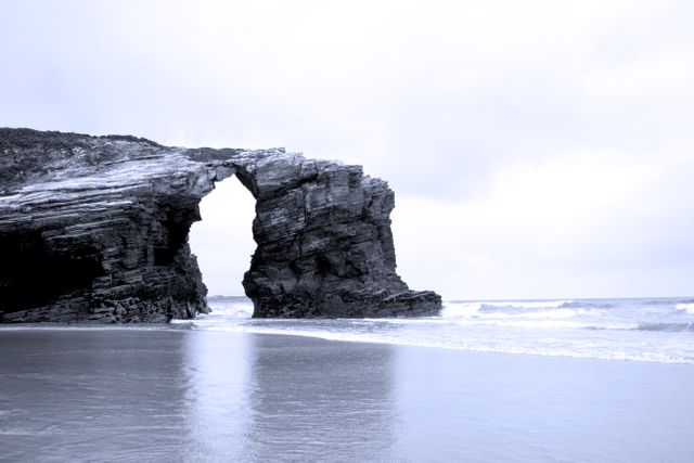 Natural rock arch formation on coast with ocean waves, under overcast sky. Perfect for illustrating coastal geography, natural landmarks, and erosion effects. Ideal for travel promotions, environmental awareness, and outdoor adventure themes.