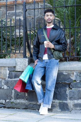 Man leaning on stone wall holding coffee and colorful shopping bags wearing leather jacket