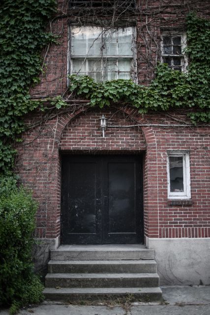 Front view of an old, abandoned red brick building with ivy covering much of the facade. The worn steps lead up to a weathered black door, showing signs of neglect and decay. The windows are dirty and panes are broken, suggesting a long period of abandonment. This type of imagery is ideal for use in projects related to urban exploration, historical reference, gothic or horror-themed materials, and settings that require an eerie or abandoned aesthetic.