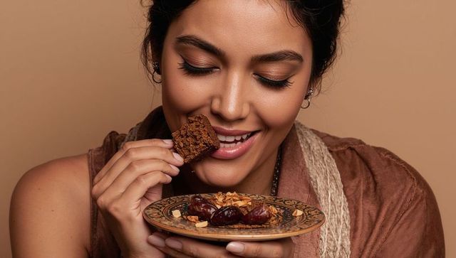 Smiling woman bringing brownie to mouth while holding ornate plate with dates and almonds