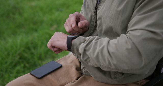 Person wearing green jacket interacting with smartwatch while sitting in park, black mobile phone placed nearby on lap. Suitable for topics on wearable technology, modern gadgets, fitness tracking, time management, convenience, and outdoor lifestyle.