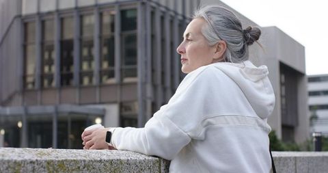 Contemplative senior woman leaning on railing in plaza wearing white hoodie and smartwatch