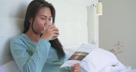Mid adult asian woman sipping water reading booklet in bed in serene minimalist bedroom