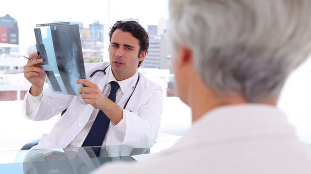 The doctor is reviewing an x-ray film in front of a patient in a bright, modern office. This video can be utilized to depict medical consultations, health check-ups, professional healthcare services, and diagnostic discussions between medical professionals and patients.