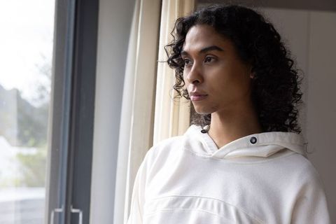 Non-binary adult standing by window wearing off-white hoodie gazing thoughtfully outdoors