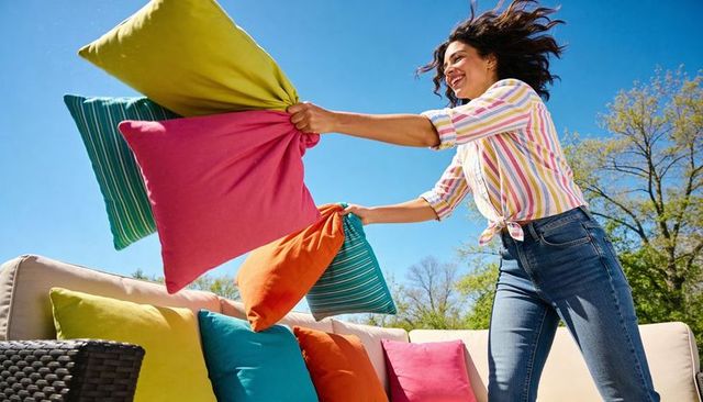 Joyful woman tossing colorful outdoor throw pillows on sunny backyard patio couch