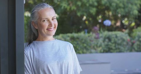 Senior woman leaning, smiling on sunny patio with lush garden backdrop
