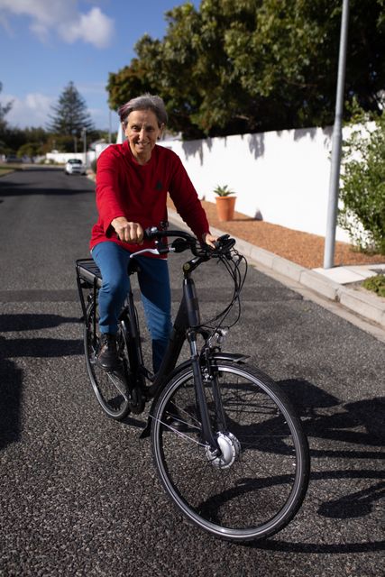 Front view  of a senior Caucasian woman with short grey hair wearing a red sweater riding a bicycle in the street, and smiling in the sun.