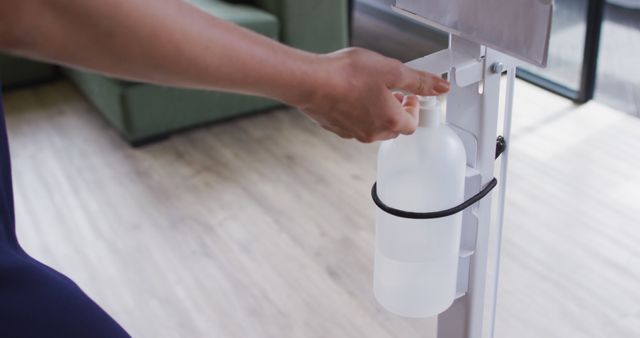 Person using hands-free hand sanitizer dispenser indoors, emphasizing hygiene and germ prevention. Useful in articles about public health measures, office cleanliness protocols, hand hygiene importance, or products for preventing the spread of illnesses.