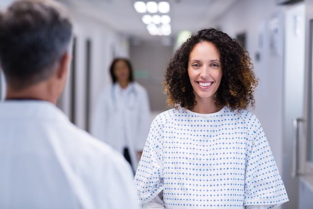 Pregnant woman standing in hospital corridor, smiling at healthcare professional. She is wearing a medical gown and has curly hair. Medical staff in the background. Ideal for use in healthcare, maternity, and hospital-related content.