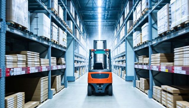 Orange forklift centered in high-bay warehouse aisle with pallet racking and led lighting