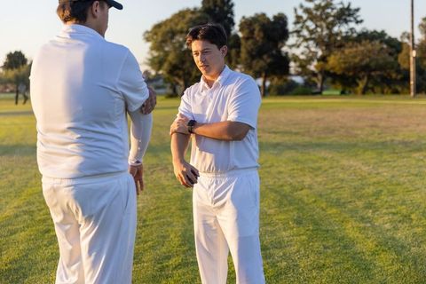 Young teammates adjusting smartwatch on cricket field during golden hour practice
