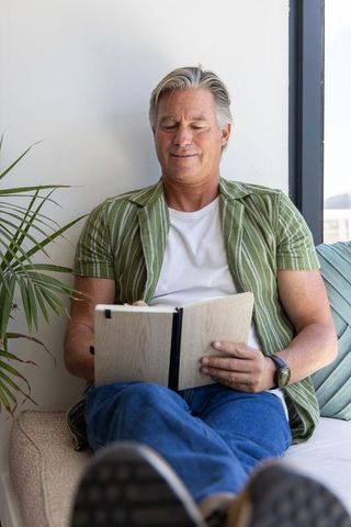 Relaxing senior man journaling on window bench in bright modern home interior, lifestyle