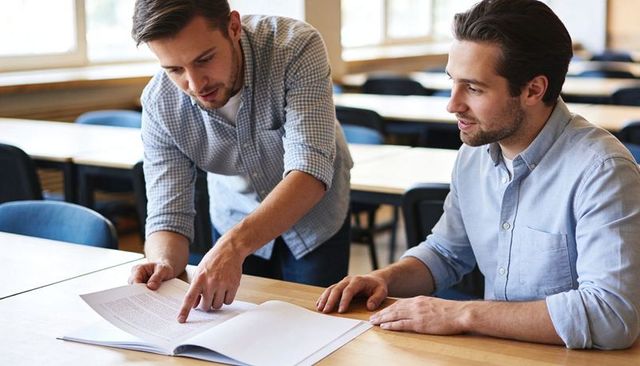 Male peers collaborating and reviewing open textbook in classroom tutoring session