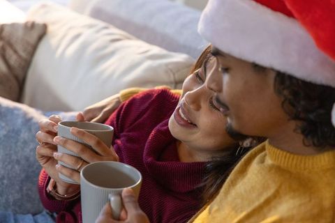 Multicultural couple sharing cozy cuddle on sofa drinking warm mugs wearing santa hat, sweaters