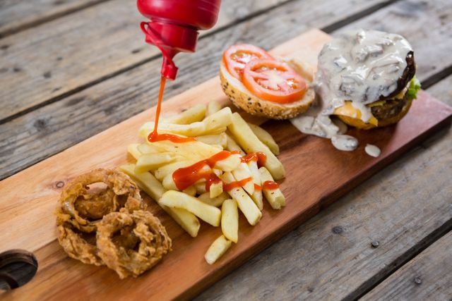 High angle view of ketchup being poured on French fries next to burger ingredients on a wooden table. The scene includes a burger bun with tomato slices, lettuce, cheese, and a creamy sauce. Onion rings are also present on the side. Ideal for use in food blogs, restaurant menus, fast food advertisements, and culinary websites.