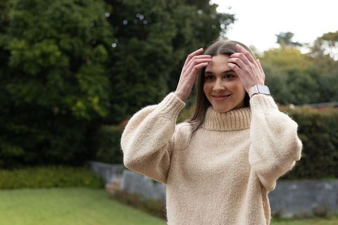 Smiling woman adjusting hair in chunky beige knit sweater wearing smartwatch in garden