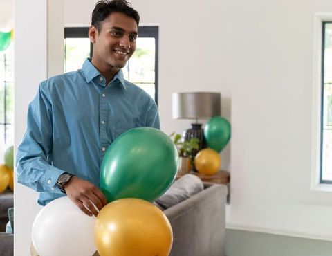 Indian man holding green gold white balloons in modern living room celebrating at home