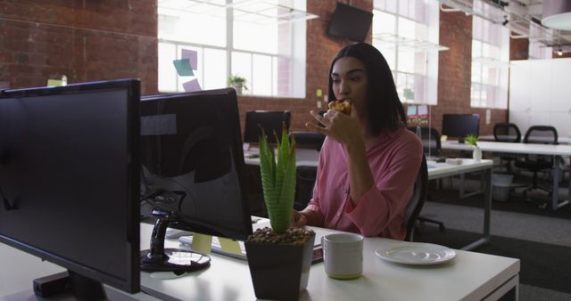 Young woman eating lunch while working on a computer in a modern office with a casual environment. Ideal for use in articles about work-life balance, office culture, productivity tips, or corporate settings.