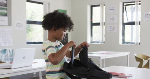 African American Boy Packing Backpack at School Desk with Laptop and Notebooks