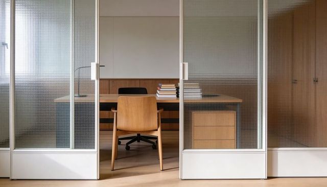 Minimalist private office with sliding textured glass doors, warm wood desk and guest chair