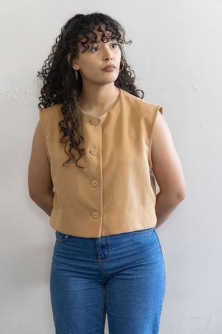 Young woman standing against textured wall wearing tan sleeveless vest and denim jeans