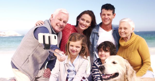 Three generations of a family, including grandparents, parents, and children, taking a selfie with a smartphone on a beach, along with their pet dog. They are smiling and enjoying their time together. Ideal for content related to family bonding, vacations, leisure activities, and multigenerational interactions.