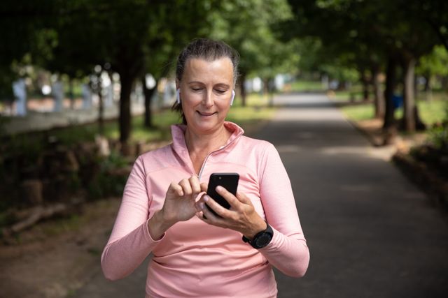 Senior woman enjoying a healthy lifestyle by exercising in the park. She is wearing sports clothes and using a smartphone with earphones, indicating she might be listening to music or a fitness app. Ideal for promoting fitness, wellness, technology use among seniors, and active retirement.