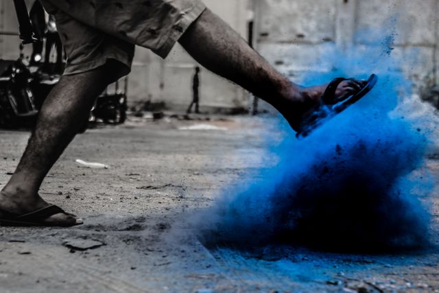This powerful shot captures a person's leg and foot in flip-flops creating a cloud of blue powder in an urban environment. Ideal for use in creative advertisements, urban-themed projects, promotional material for festivals or events, and artistic designs conveying energy and motion.